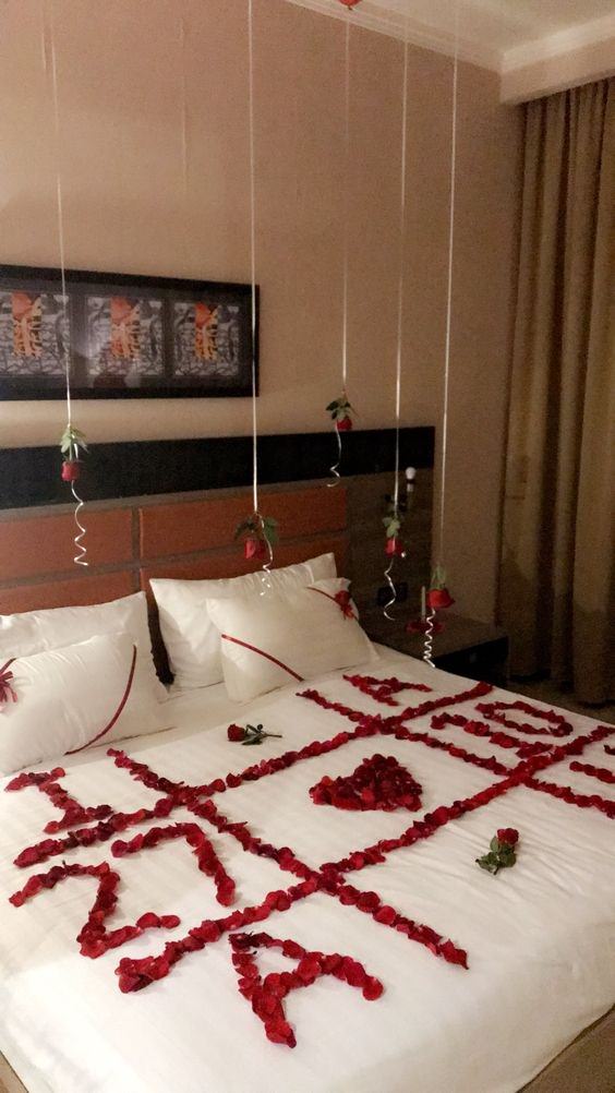 اتاق عروسی که شاخه های گل رز توسط بادکنک های هلیومی از آن آویزان شده است و روی روتختی سفید آن گلبرگ های قرمز به شکل قلب، حروف و اعداد قرار داده شده است